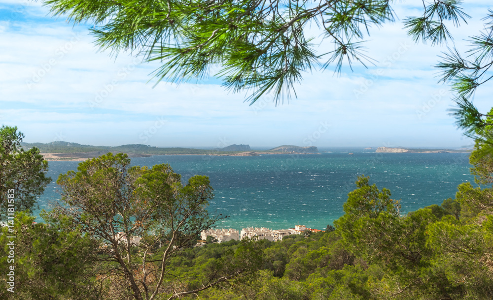 Wide angle view through trees from high point on nearby hillside out to see and the familiar islands just offshore of Ibiza.