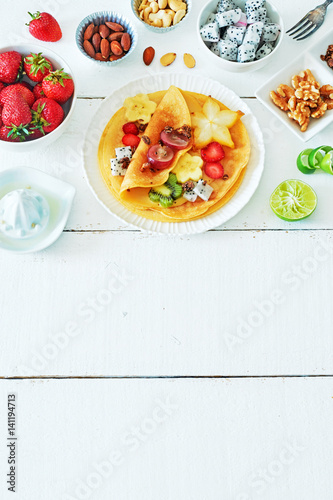 Vertical top view of healthy breakfast with crepes and exotic fruits. Thin pancakes with starfruit  dragon fruit  kiwi  banana  strawberries  walnut  almond and lime on a white table. Copy space.