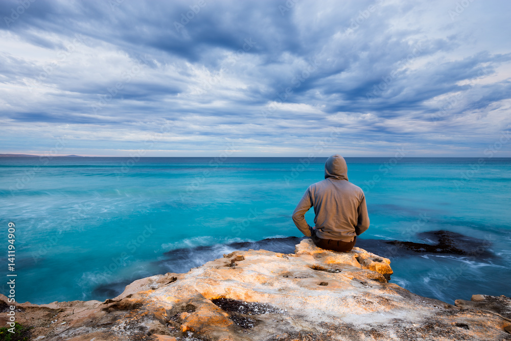 A man sits on the edge of a rugged limestone cliff and looks over a stormy ocean view in South Australia.