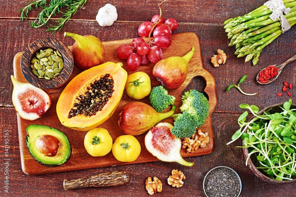 Vegetarian ingredients on rustic board over wooden texture background. Top view of papaya, figs, grape, tomato, avocado on rustic board, sprouts, chia and pumpkin seeds, goji. 