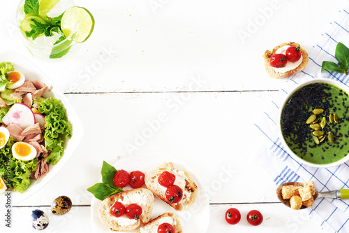 Top view of healthy lunch or dinner meal on a white table with copy space. Green puree soup, tuna salad with potato, lettuce and quail egg, bruschetta with roasted tomato, mint & lime lemonade.