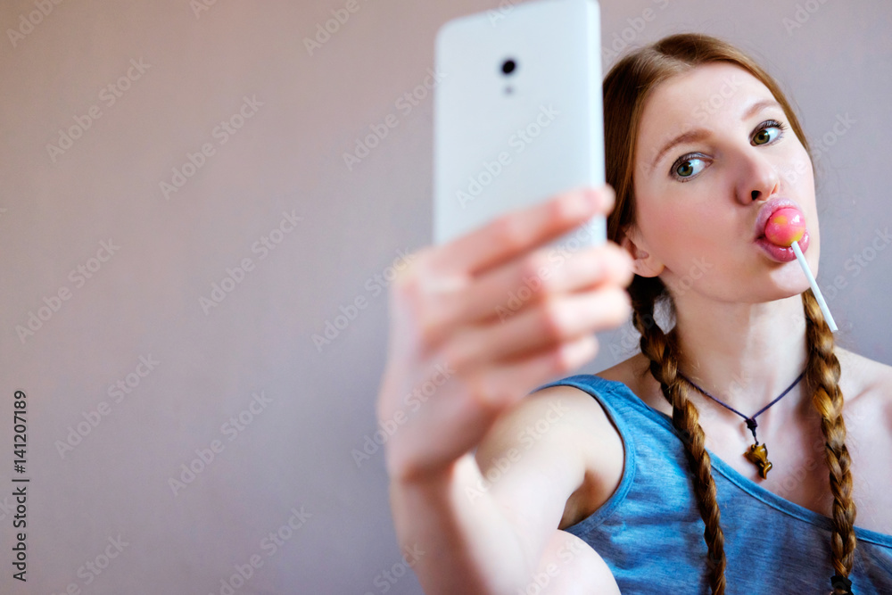 Redhead girl is licking a lollipop and making a selfie at smartphone. Redhead girl is licking a lollipop and making a selfie at smartphone.