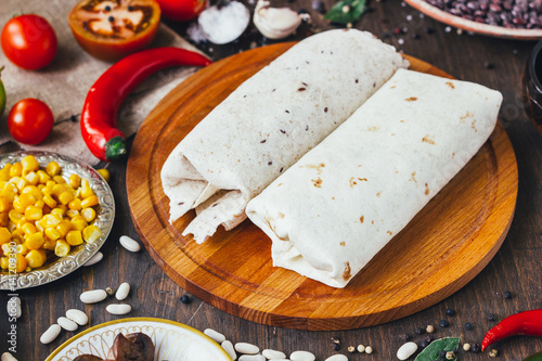 Vegetarian burrito on wooden board over black table surrounded by ingredients.