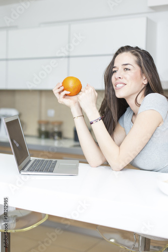 Morning scene. Beautiful woman holding orange in front of laptop