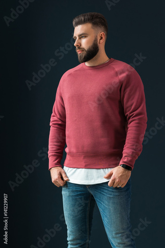 Fotografija A young bearded handsome male hipster, dressed in a red pullover with long sleeves and jeans, stands indoors