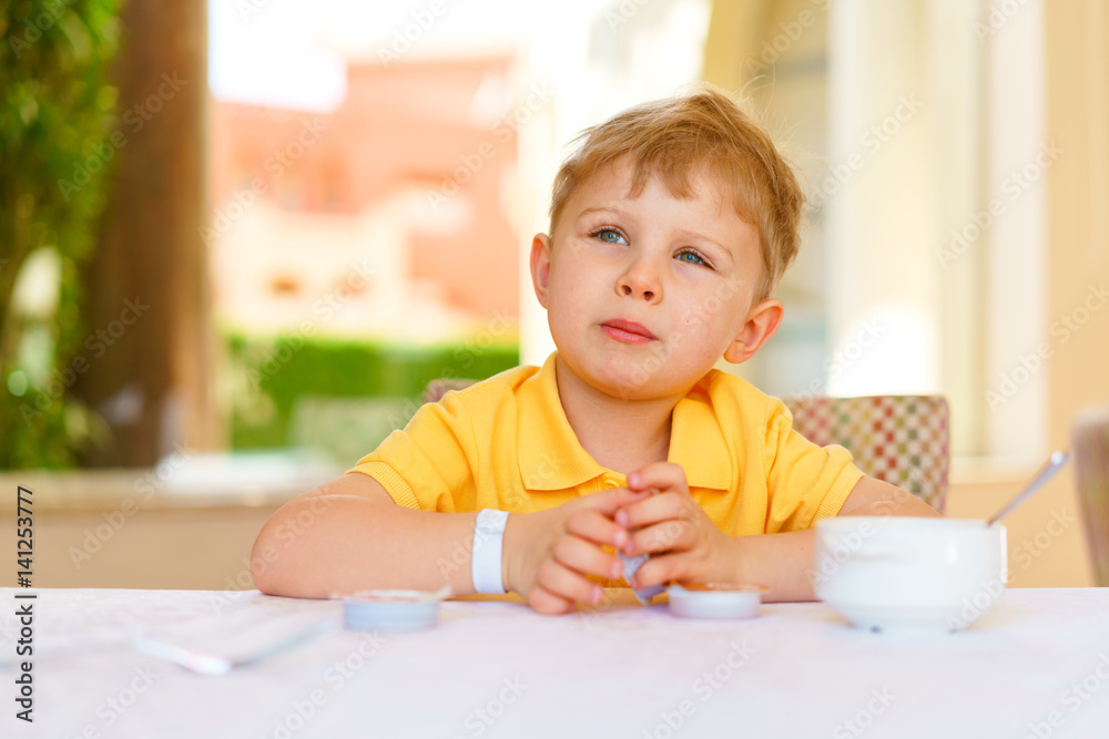 Kid looks dreamily into the distance, sitting at a table with breakfast in a summer terrace lit with daylight. Boy dressed in an casual yellow polo. Kid looks dreamily into the distance, sitting at a table with breakfast in a summer terrace lit with daylight. Boy dressed in an casual yellow polo.