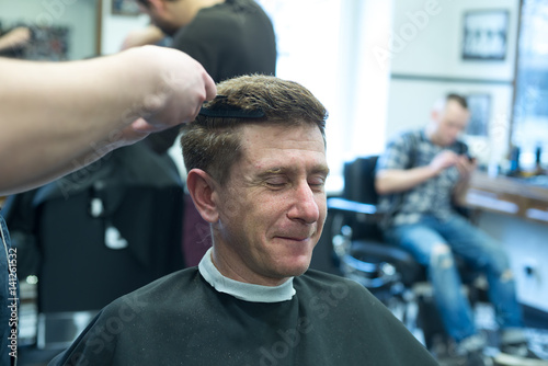 Smiling man, client in a barbershop, closing his eyes, enjoys the process