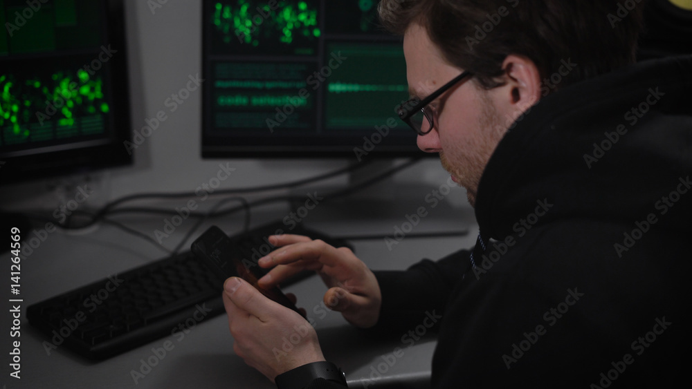The underground organization. Two computers and a laptop on desk. A man in a dark jacket and glasses looks at the article on your smartphone and tries to understand the information The underground organization. Two computers and a laptop on desk. A man in a dark jacket and glasses looks at the article on your smartphone and tries to understand the information