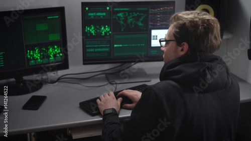 Experienced hacker carries out cyber attacks on major companies server to steal money from their accounts. Man with glasses at the same time work for multiple computers in an illegal secret office. © kustvideo