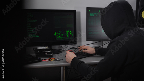 Back view of a hooded hacker making cyber attack on bank network. Man in black is typing fast on keyboard and stealing data of credit cards with the process viewed on screens in front of him.