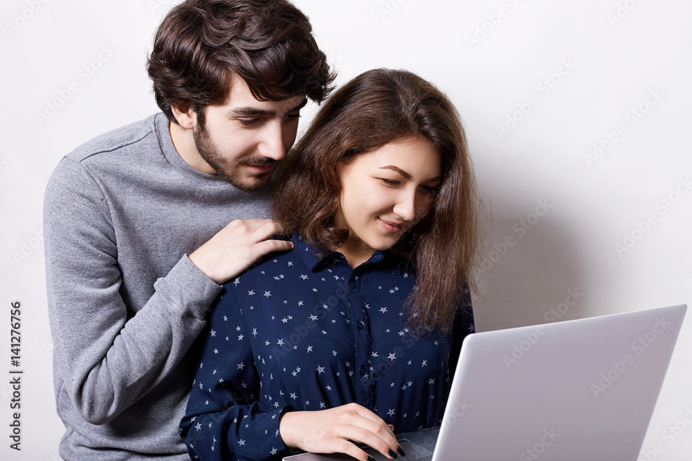 Young couple using laptop computer together. Attractive bearded man dressed in casual sweater holding his hand on his girlfriend`s shoulder looking into the screen while having video call. Young couple using laptop computer together. Attractive bearded man dressed in casual sweater holding his hand on his girlfriend`s shoulder looking into the screen while having video call.