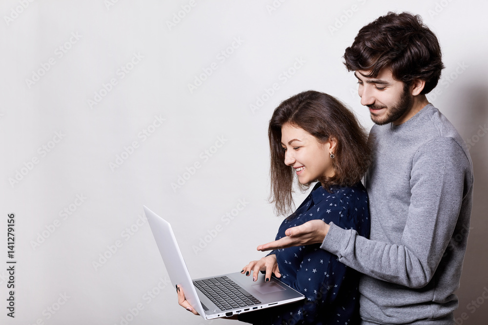 A sideways portrait of beautiful couple watching videos online standing close to each other holding laptop using free wireless internet connection, smiling, looking at screen. People and lifestyle.