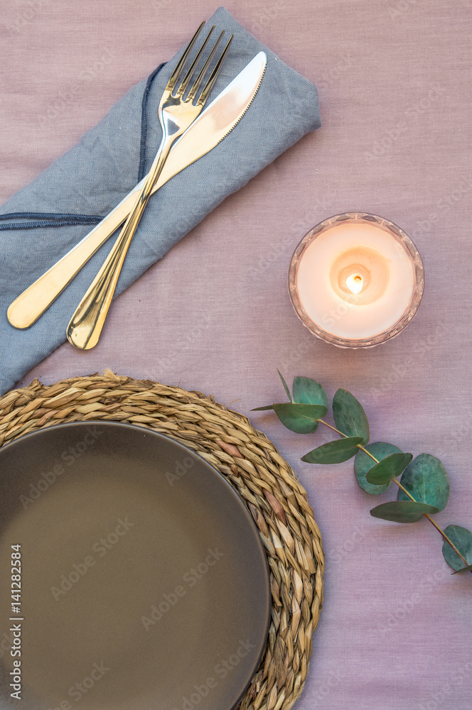 Elegant table setting on purple linen cloth dark plate rattan coaster cutlery napkin, burning candle, twig of silver dollar eucalyptus, top view
