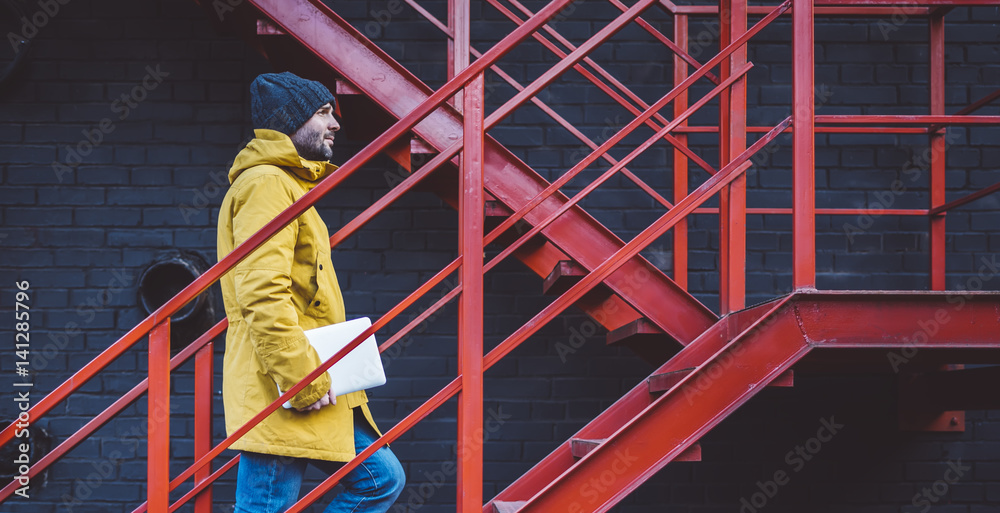 Hipster with yellow backpack, jacket rises on red retro ladder with computer in hands, freelance holding laptop in spring street outdoor, tourist man traveler connect internet, process in workspace