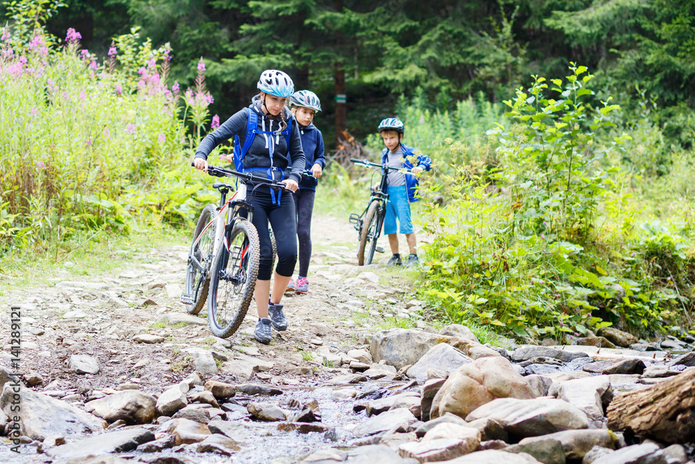 Fototapeta premium Young woman with her children on bikes wade the mountain creek