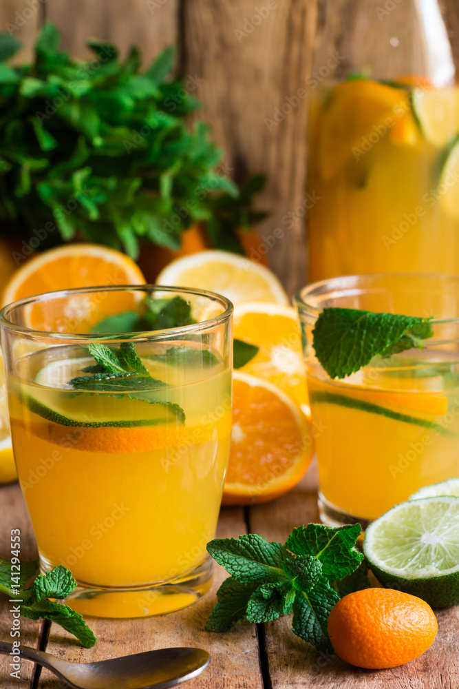 Refreshing citrus lemonade from lime oranges fresh mint in glasses and bottle, sliced scattered fruits, wood kitchen table by window Refreshing citrus lemonade from lime oranges fresh mint in glasses and bottle, sliced scattered fruits, wood kitchen table by window