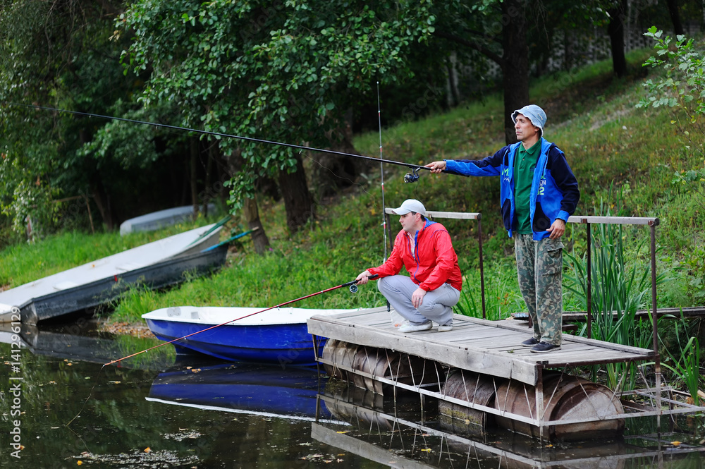 Two fishermen with fishing rods catching fish in the river standing on the pier bridge Two fishermen with fishing rods catching fish in the river standing on the pier bridge