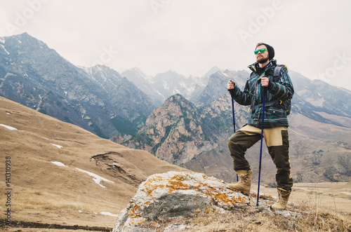 Tired bearded hipster with binoculars in his hands sits on a stone among the mountains and looks out into the distance
