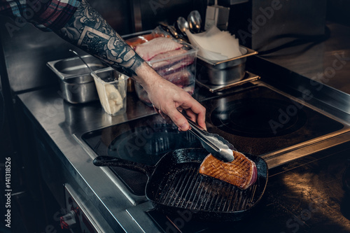 A man with tattooed arm grilling a beef in a kitchen.