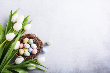 Beautiful white tulips with colorful quail eggs in nest on light gray stone background. Spring and Easter holiday concept with copy space.