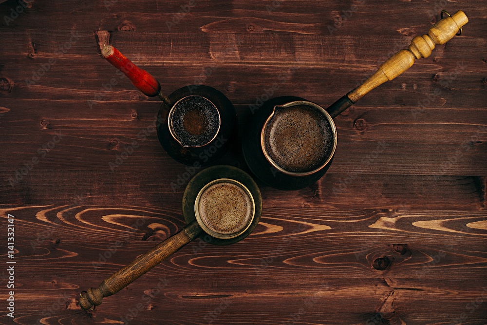 Hot coffee in shabby turkish pots cezve with tasty crema  on brown old wooden board background, top view. Rustic style.