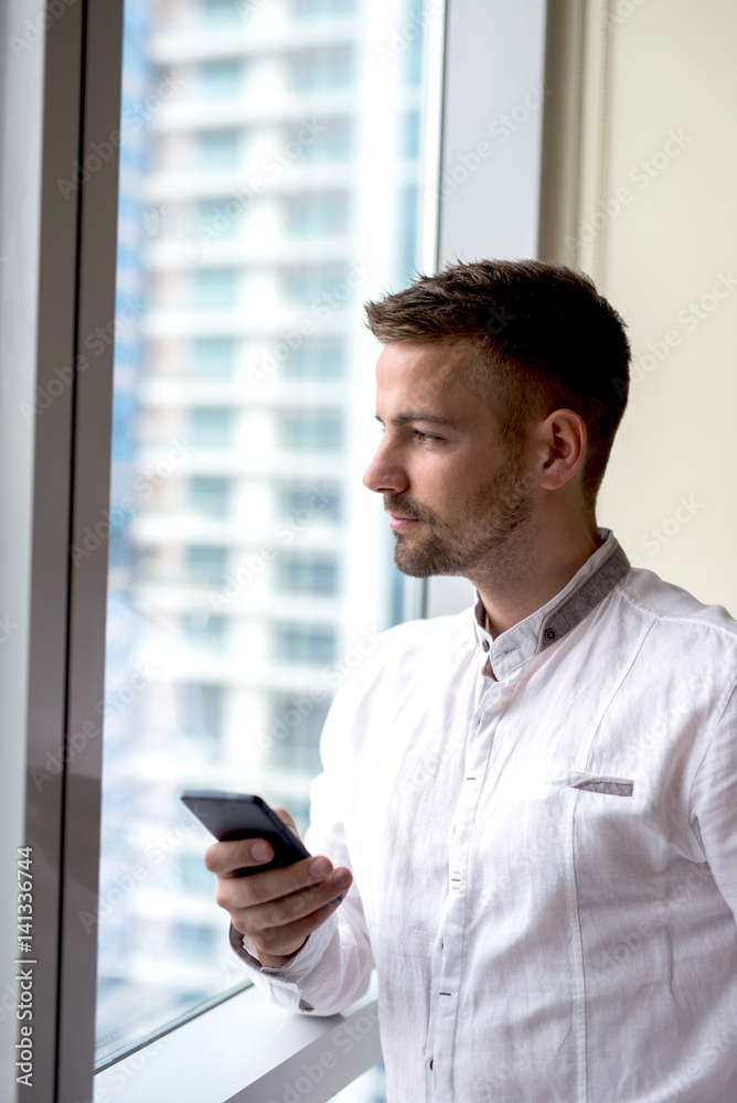 Man standing next to bright window holding phone, looking away. Man standing next to bright window holding phone, looking away.