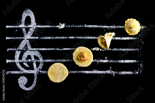Fototapeta Naklejka Na Ścianę i Meble -  Ravioli - cooking music. Dough with minced meat on a black background and a treble clef from flour