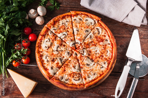 Traditional italian cuisine. Appetizing fresh pizza with mushrooms served on wooden background with ingredients and special knife, flat lay.