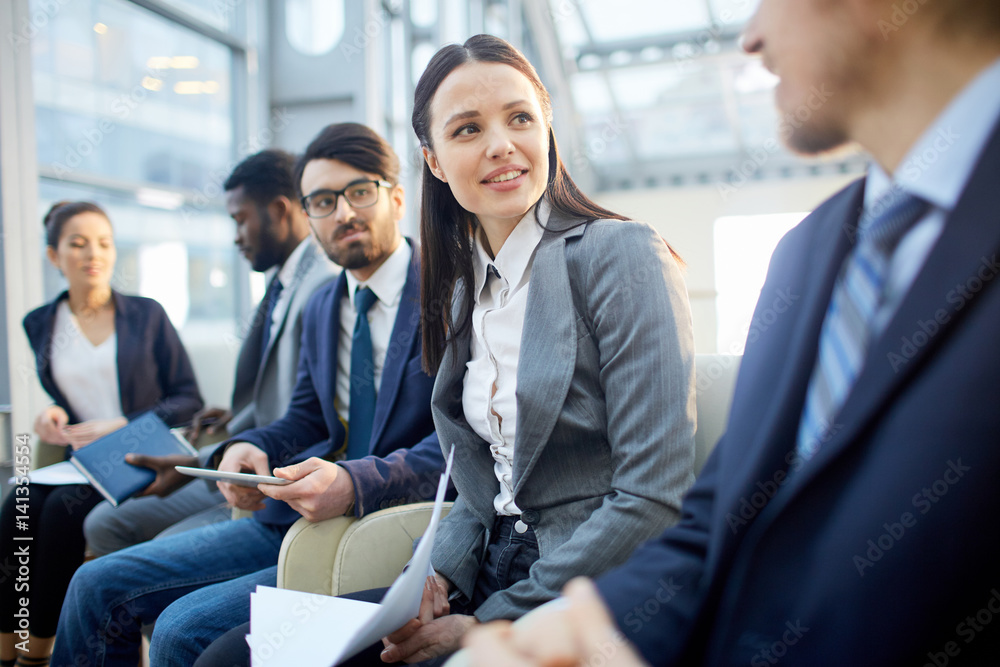 Multi-ethnic group of business people sitting in row in modern glass hall, focus on young beautiful brunette woman talking to unrecognizable businessman Multi-ethnic group of business people sitting in row in modern glass hall, focus on young beautiful brunette woman talking to unrecognizable businessman