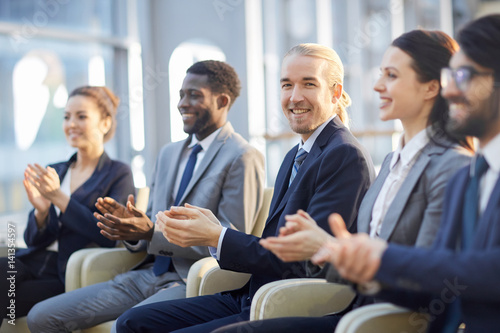 Multi-ethnic group of smiling business people sitting in row in modern glass hall and clapping, focus on young cheerful  businessman in center