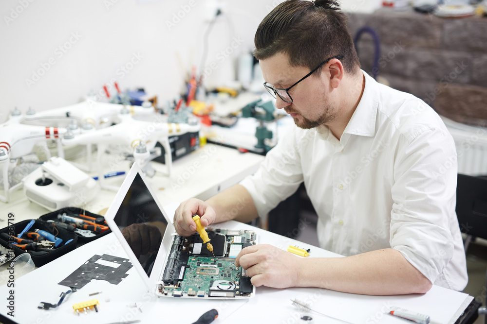 Portrait of modern man wearing glasses busy with electronics in workshop: disassembling laptop and reconstructing it with different tools and parts on table