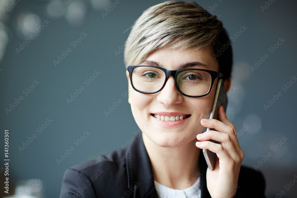Head and shoulders portrait of smiling businesswoman in stylish eyeglasses looking at camera while calling to her colleague on smartphone Head and shoulders portrait of smiling businesswoman in stylish eyeglasses looking at camera while calling to her colleague on smartphone