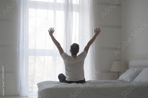 Fototapeta Naklejka Na Ścianę i Meble -  Happy young man stretching in bed after waking up, back view