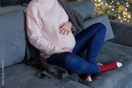 Pregnant girl sits on a sofa near a Christmas tree and a fireplace