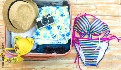 Open the suitcase with tourist things: women's hat, swimsuit, camera, denim shorts, dresses, sunglasses, perfumes, nail polish, mobile phone, tablet on wooden background