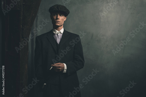 Retro 1920s english gangster standing with cigarette. Wearing suit and flat cap.