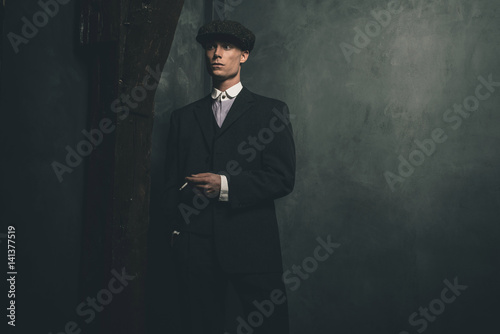 Retro 1920s english gangster standing with cigarette. Wearing suit and flat cap.