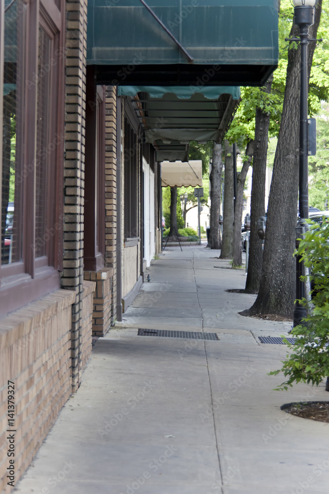 View of empty cement sidewalk leading through trees and various shops.
