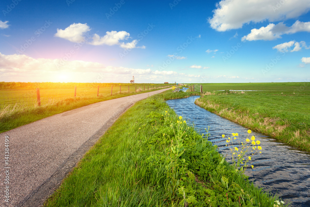 Landscape with green grass field, road, lighthouse, river and blue sky with clouds reflected in water at amazing sunset in spring. Colorful nature background. Agriculture. Green meadow. Water canal