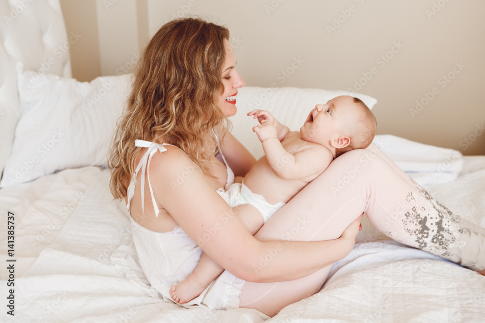 Group portrait of beautiful middle age white Caucasian mother and baby sitting together on bed in bedroom talking playing laughing, early development Group portrait of beautiful middle age white Caucasian mother and baby sitting together on bed in bedroom talking playing laughing, early development