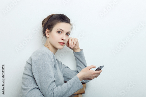 Indoor close up portrait of charming young lady of European appearance wearing grey sweatshirt sitting browsing on smartphone against white wall background. Emotions and facial expression concept