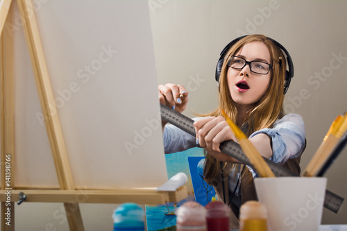 The blonde listens to music and draws. Student of art school. A woman paints a painting on canvas. A blonde in glasses uses a ruler.