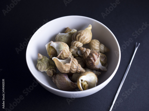 Whelks (sea snails) in white bowl with snail fork isolated on black background