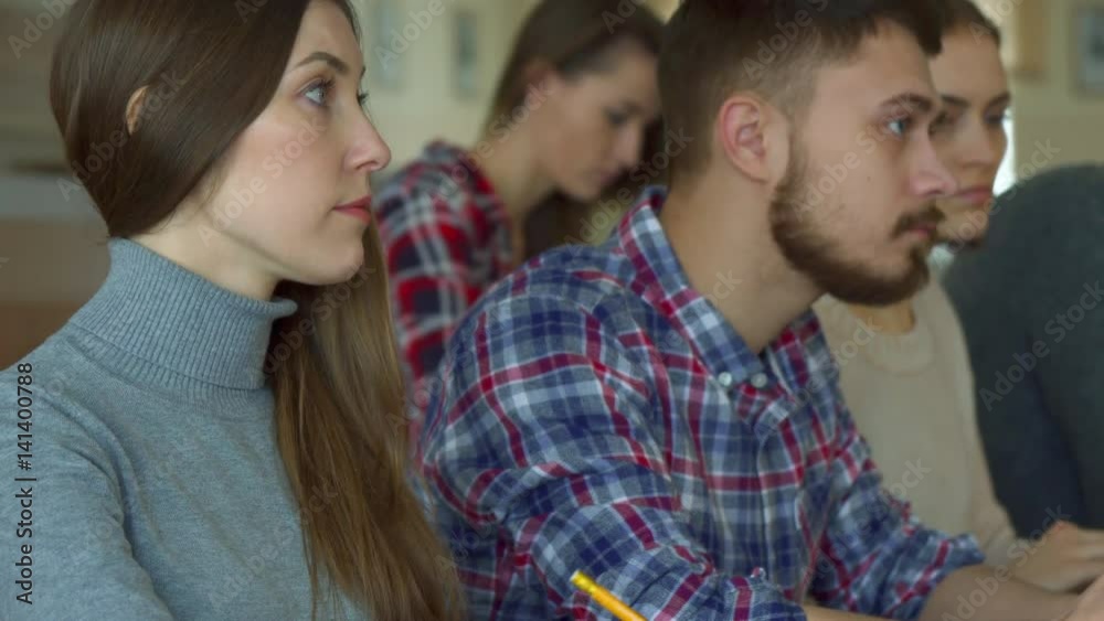 Young caucasian people studying at the college. Close up of pretty brunette girl smiling for the camera at the lecture hall. Attractive female student sitting against background of her bearded male Young caucasian people studying at the college. Close up of pretty brunette girl smiling for the camera at the lecture hall. Attractive female student sitting against background of her bearded male