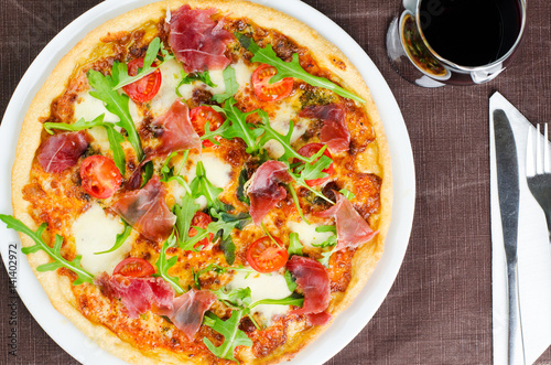 Tasty Italian Pizza With Prosciutto, Fresh Tomatoes And Rucola Next To Glass Of Red Wine