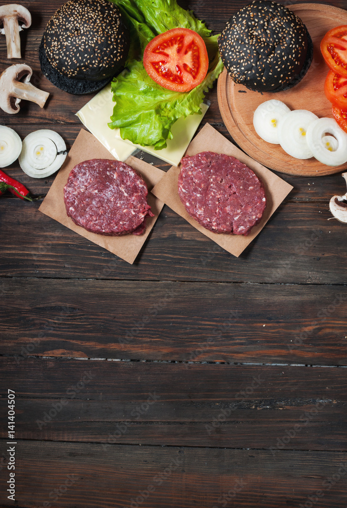 Homemade hamburger ingredients. Raw minced beef, fresh black bun, slice of cheese, tomato, onion rings, lettuce on wood background