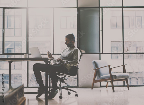 Bearded coworker man relaxing in chair at modern coworking studio and using smartphone.Panoramic windows on blurred background.Horizontal.