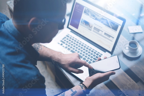 Businessman pointing finger on smartphone screen.Adult tattooed coworker in eyeglasses working at sunny office on laptop.Blurred background.Horizontal.