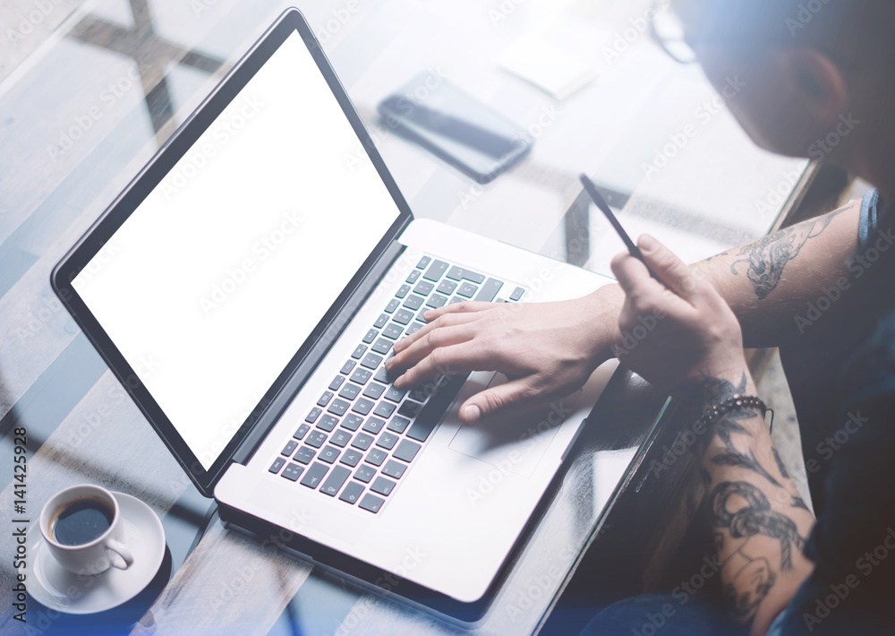 Fototapeta premium Adult tattooed businessman working on mobile computer at sunny office.Businessman typing on notebook keyboard.White clean laptop screen.Horizontal mockup,blurred background.