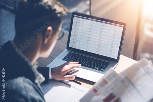 Closeup view of banking finance analyst in eyeglasses working at sunny office on laptop while sitting at wooden table.Businessman analyze stock report on notebook screen.Blurred background,horizontal. photo